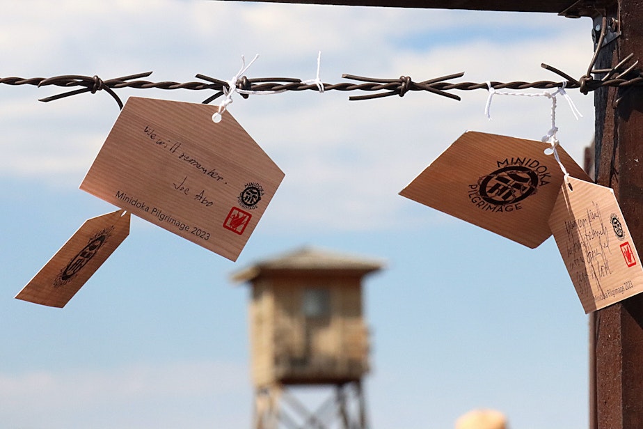 caption: Emas with messages of hope and honoring loved ones hang off of mock barbed wire as the replica guard tower stands at the entrance of the prison camp.