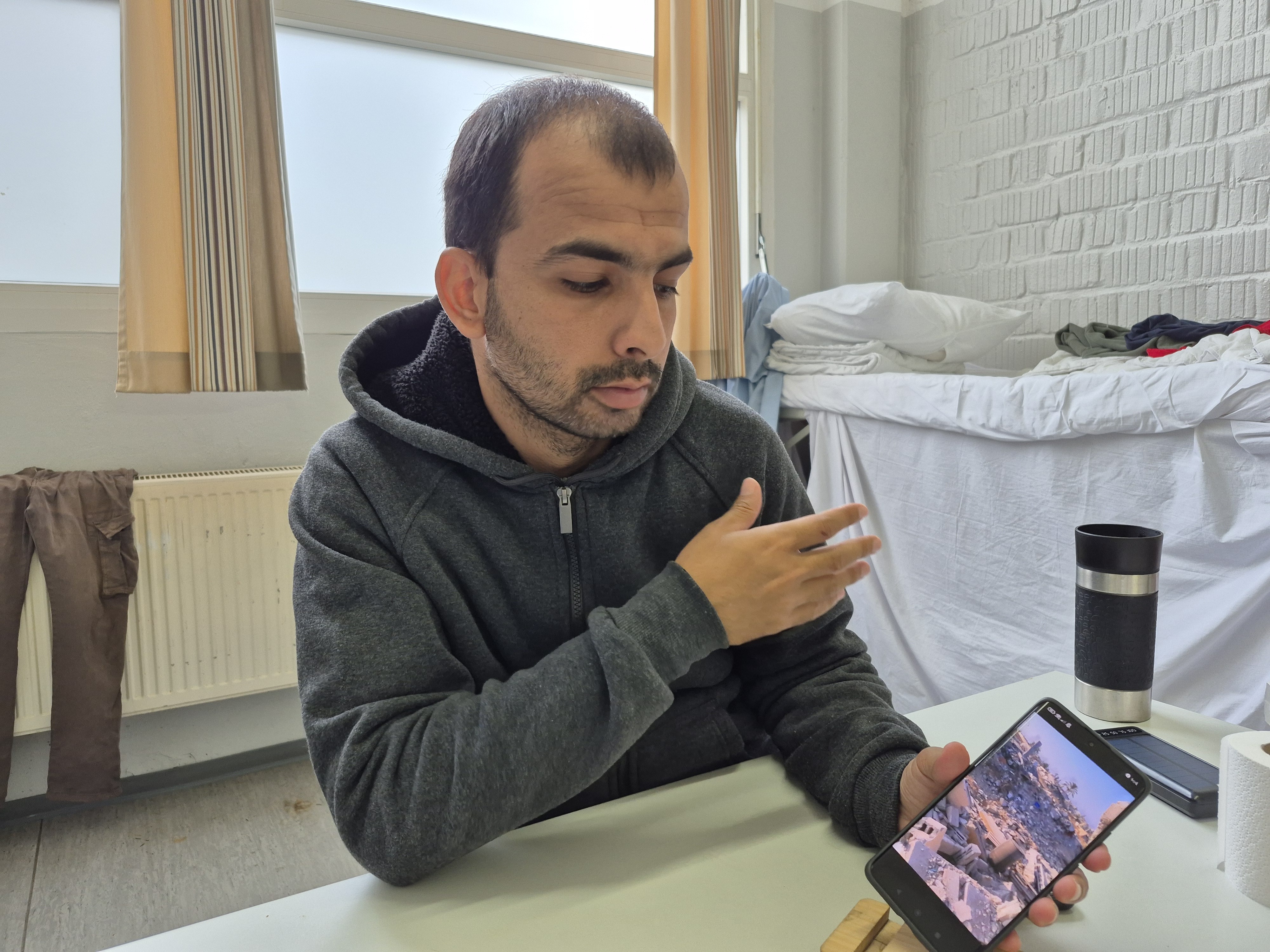 caption: Mohammed Abu Daqqa, a 31-year-old Palestinian who left Gaza, scrolls through images on his phone at a refugee welcome center in Germany. He describes how he rode a jet ski across the Mediterranean to take refuge in Europe, and is determined to get his family out of Gaza.