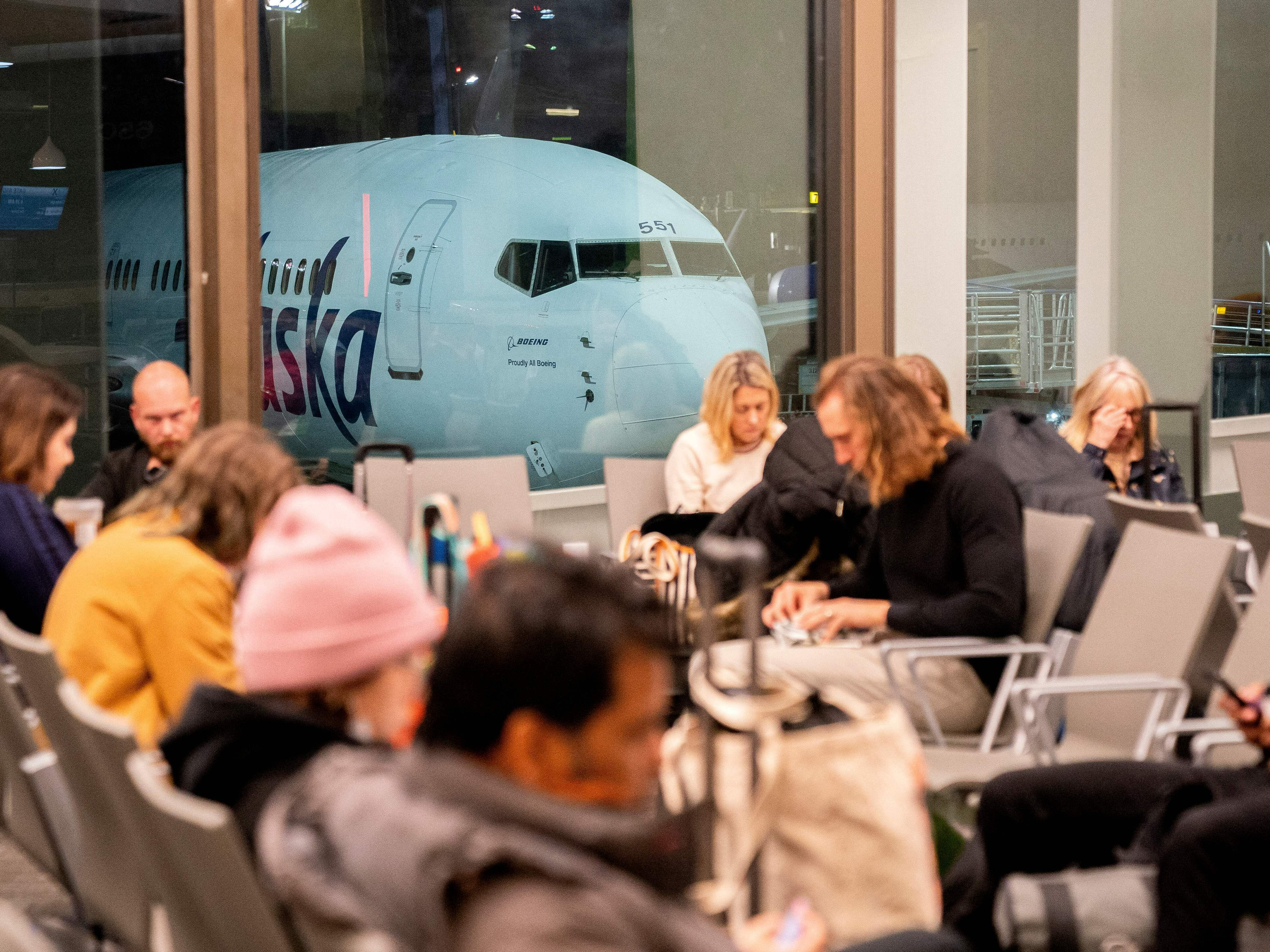 caption: Travelers wait in the terminal as an Alaska Airlines plane sits at a gate at Los Angeles International Airport early Thursday.