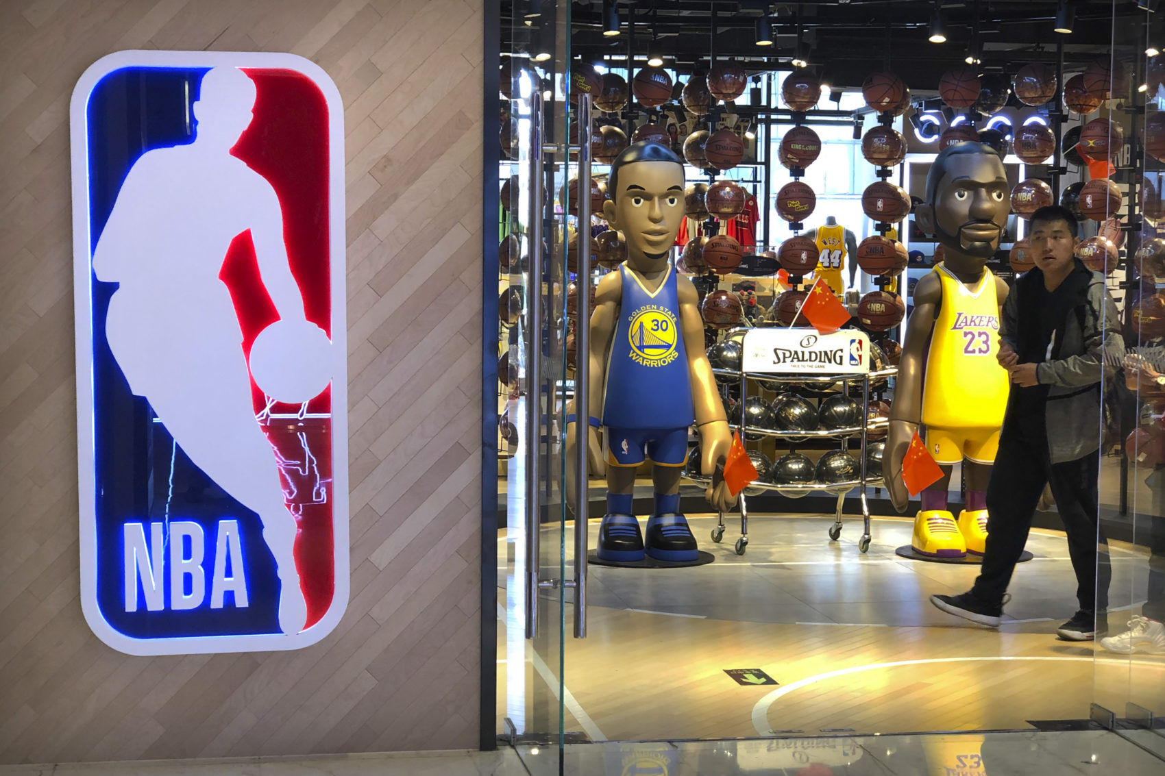 caption: A man walks past statues of NBA players Stephen Curry of the Golden State Warriors, left, and Lebron James of the Los Angeles Lakers holding Chinese flags in the entrance of an NBA merchandise store in Beijing, Tuesday, Oct. 8, 2019. (Mark Schiefelbein/AP)