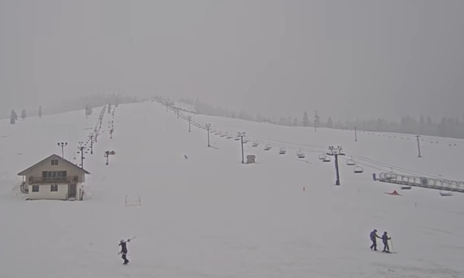 caption: Several skiers cross in front of the webcam view at the snowy summit west base area at The Summit at Snoqualmie on March 10, 2026.