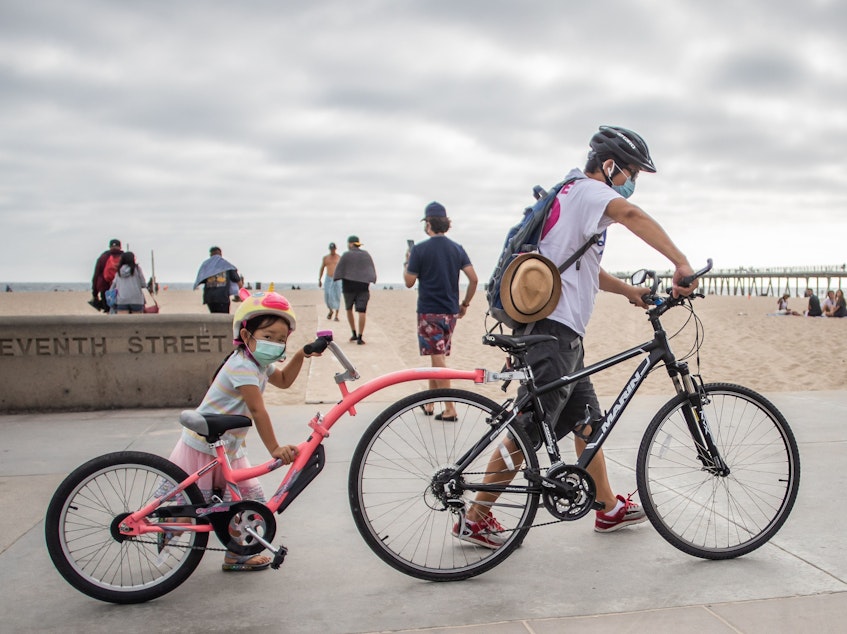 caption: A girl and her father wear facemasks while they push their bikes last summer in Hermosa Beach, Los Angeles. There aren't yet coronavirus vaccines approved in the U.S. for kids under 12 — which means they should keep masking, according to the CDC.