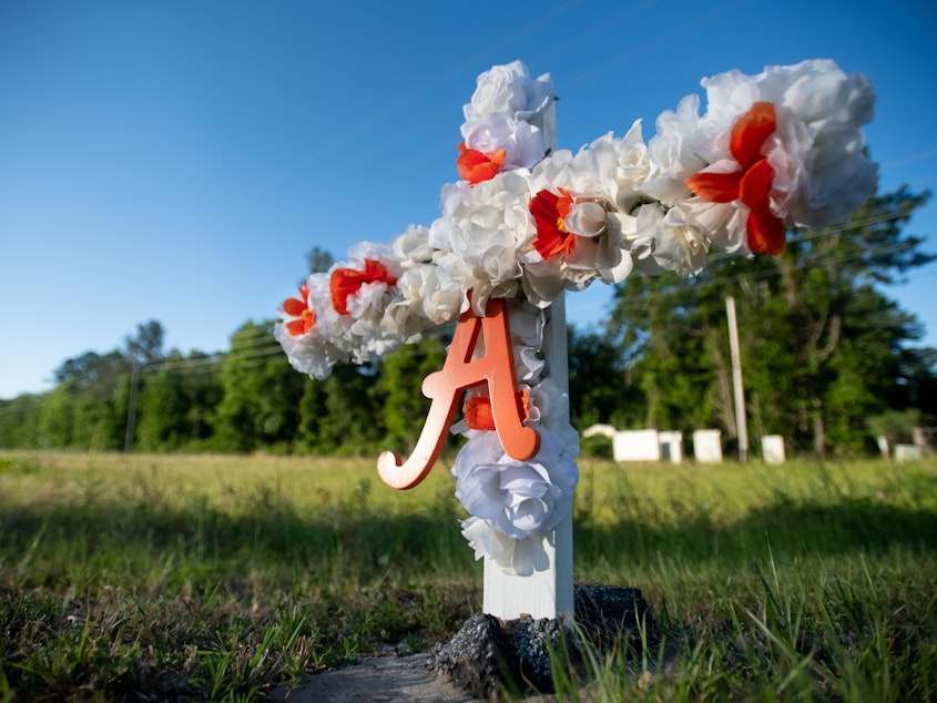 caption: A cross with flowers and the letter "A" sits at the entrance to the Satilla Shores neighborhood where Ahmaud Arbery was shot and killed.
