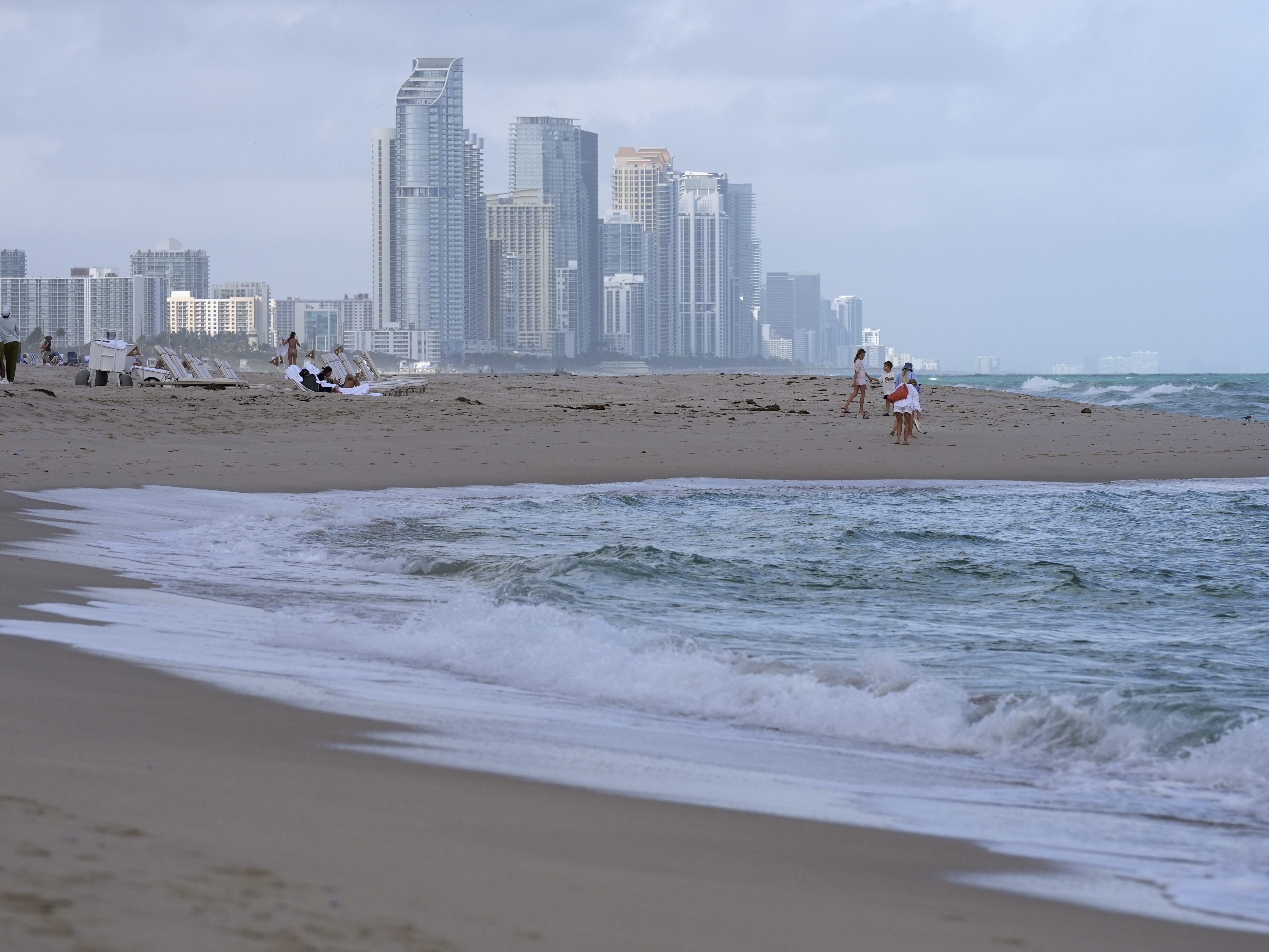 caption: People walk along the beach in Surfside, Fla., near the skyline of Sunny Isles Beach, on Dec. 17, 2024.