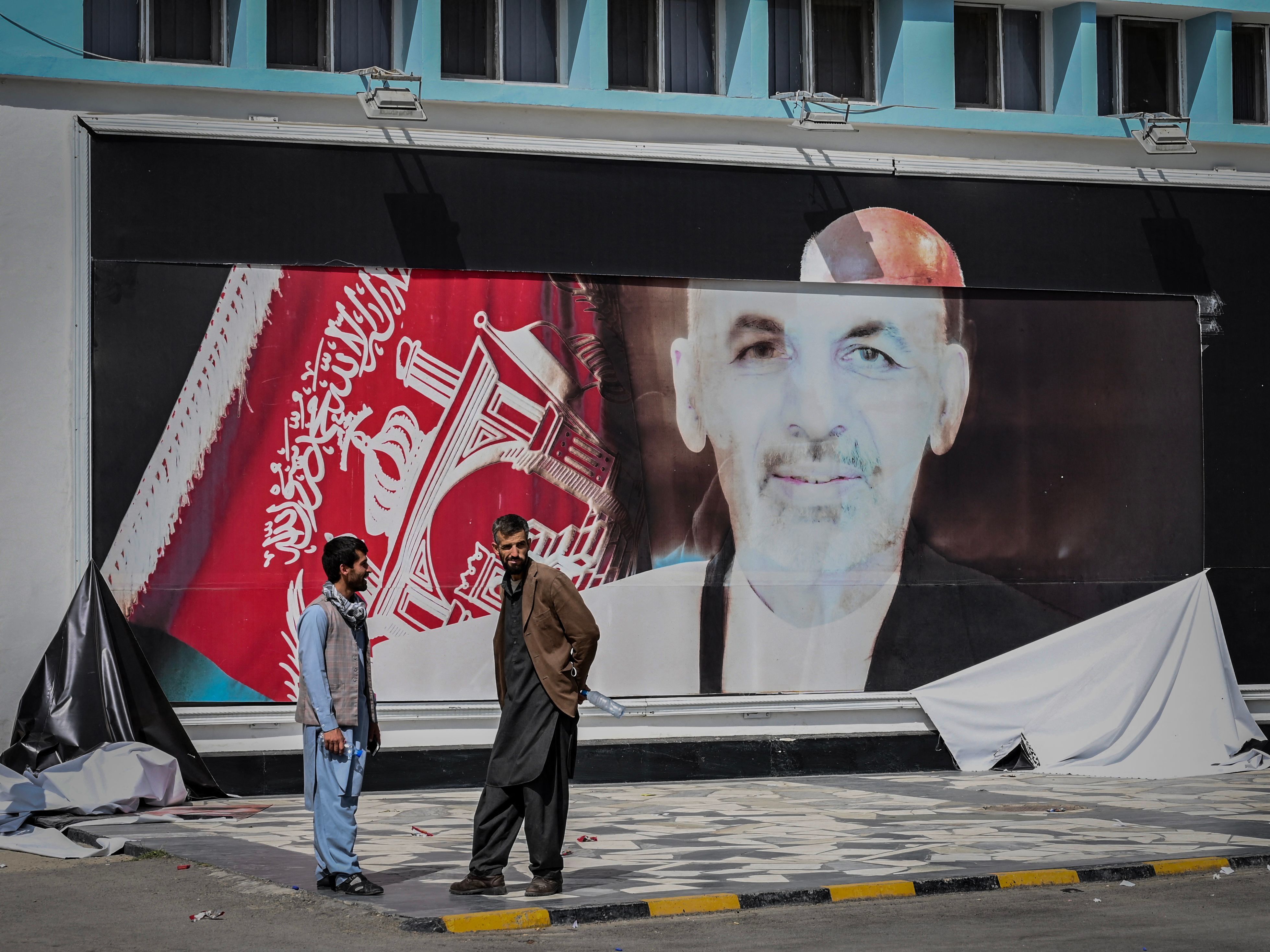 caption: Men stand next to a torn poster of ousted Afghan President Ashraf Ghani at the Kabul airport on Monday. Ghani fled Afghanistan over the weekend as Taliban forces closed in on Kabul.