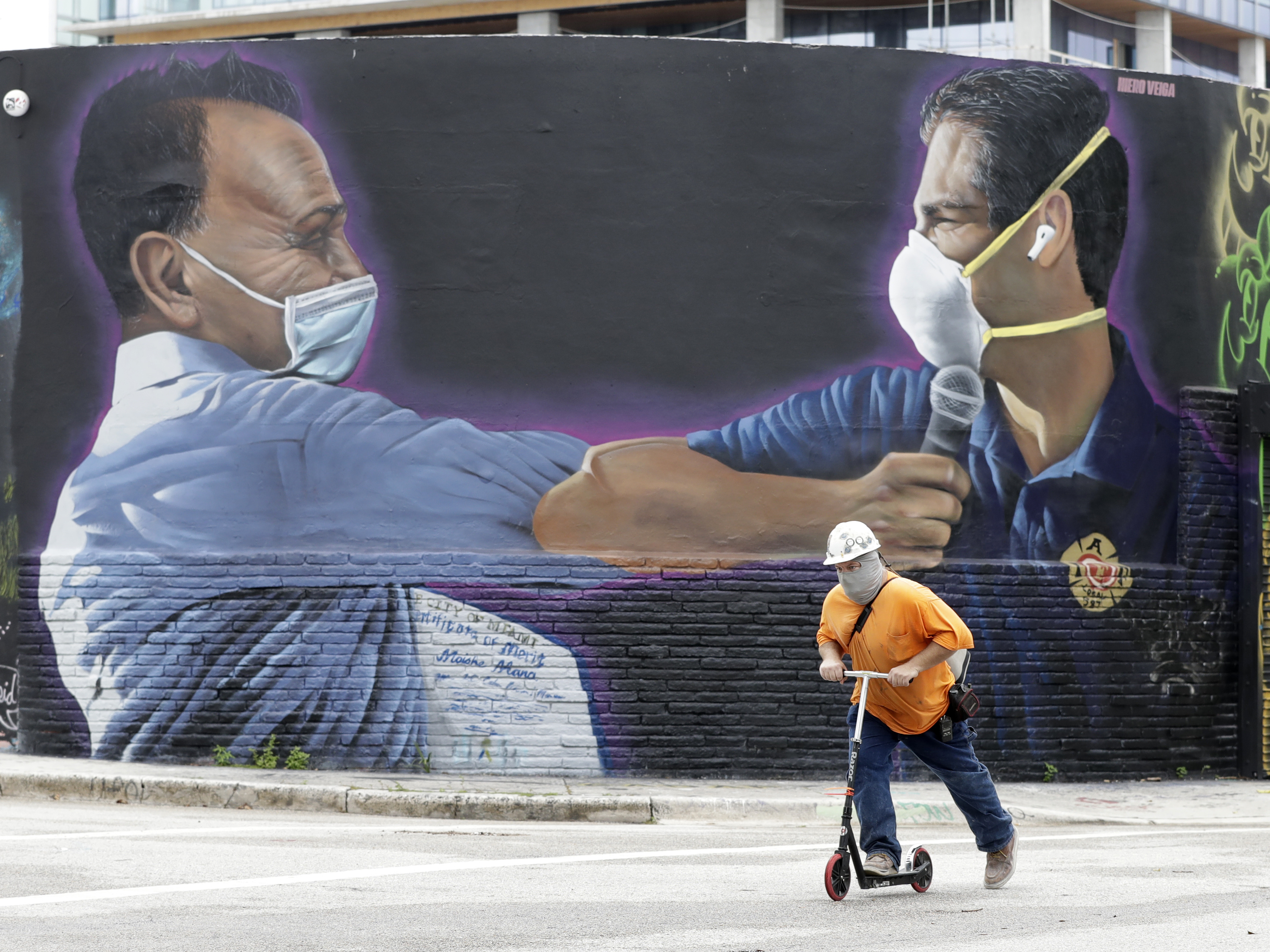 caption: A construction worker rides a scooter Monday in Miami past a Hiero Veiga mural of businessman Moishe Mana (left) and Miami Mayor Francis X. Suarez wearing masks.