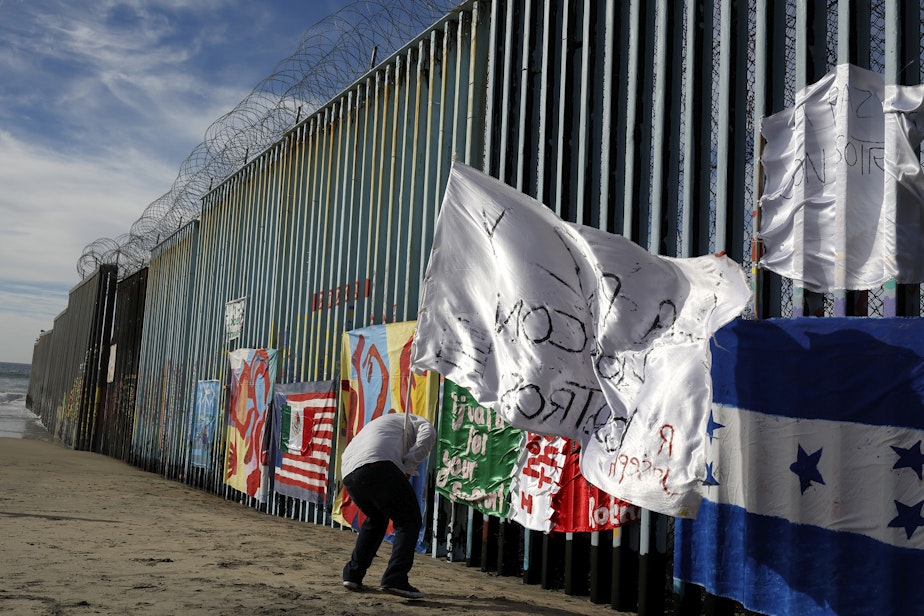 caption: Joseph, an migrant from Honduras, plants a white flag with the words, "peace and God with us," in front of the border wall during an art display on the border wall, topped with razor wire, Tuesday, Jan. 8, 2019, on the beach in Tijuana, Mexico. (Gregory Bull/AP)