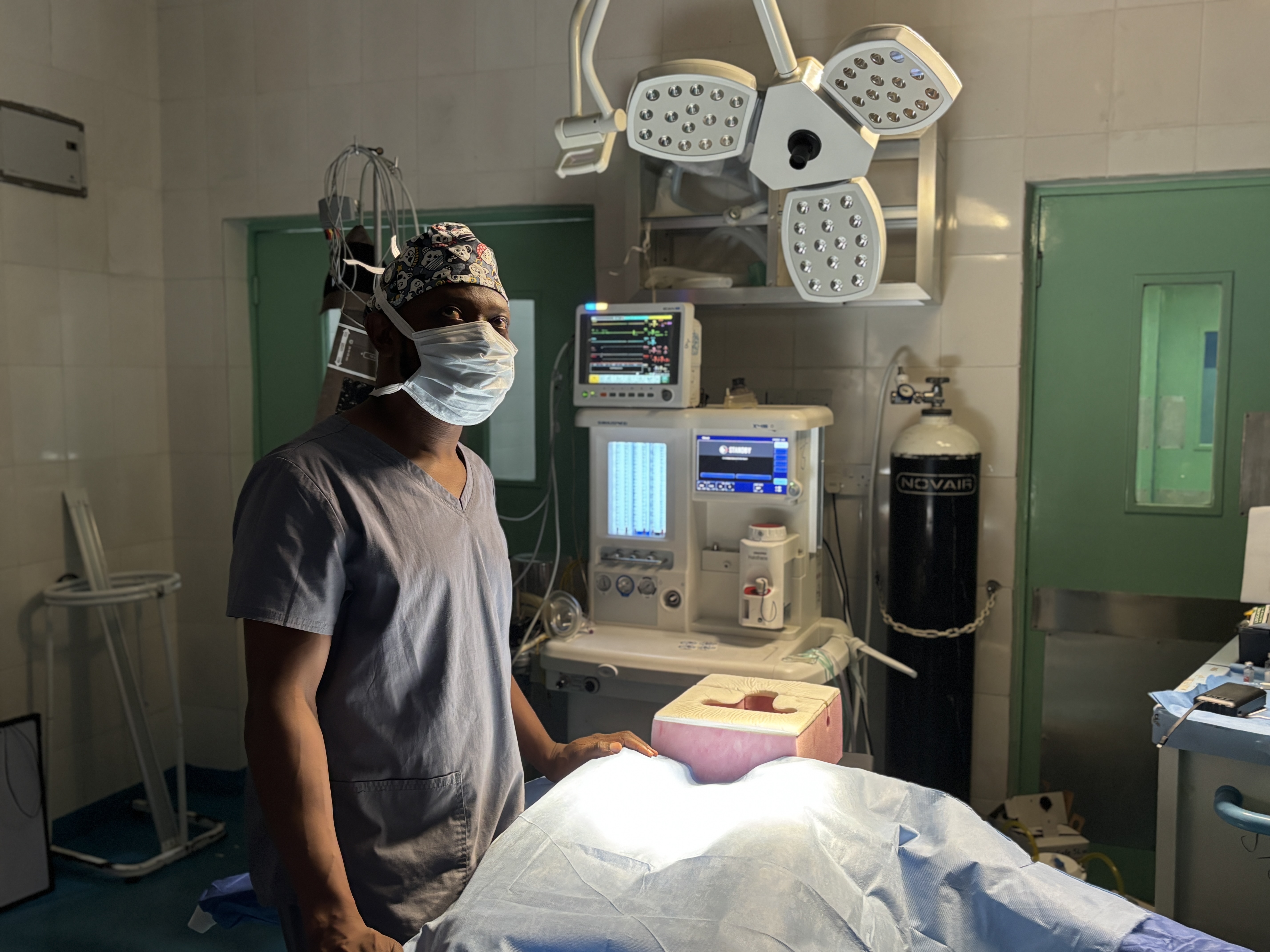 caption: Dr. Alieu Kamara of Sierra Leone stands in front of a surgical bed outfitted with improvised bolsters and a foam headrest. He is the country's first and only neurosurgeon. "We used to lose a lot of patients due to head injuries, spinal cord injuries, spine fractures and the like," Kamara said. "There was nothing we could do for them." That reality inspired him to pursue training in the field.