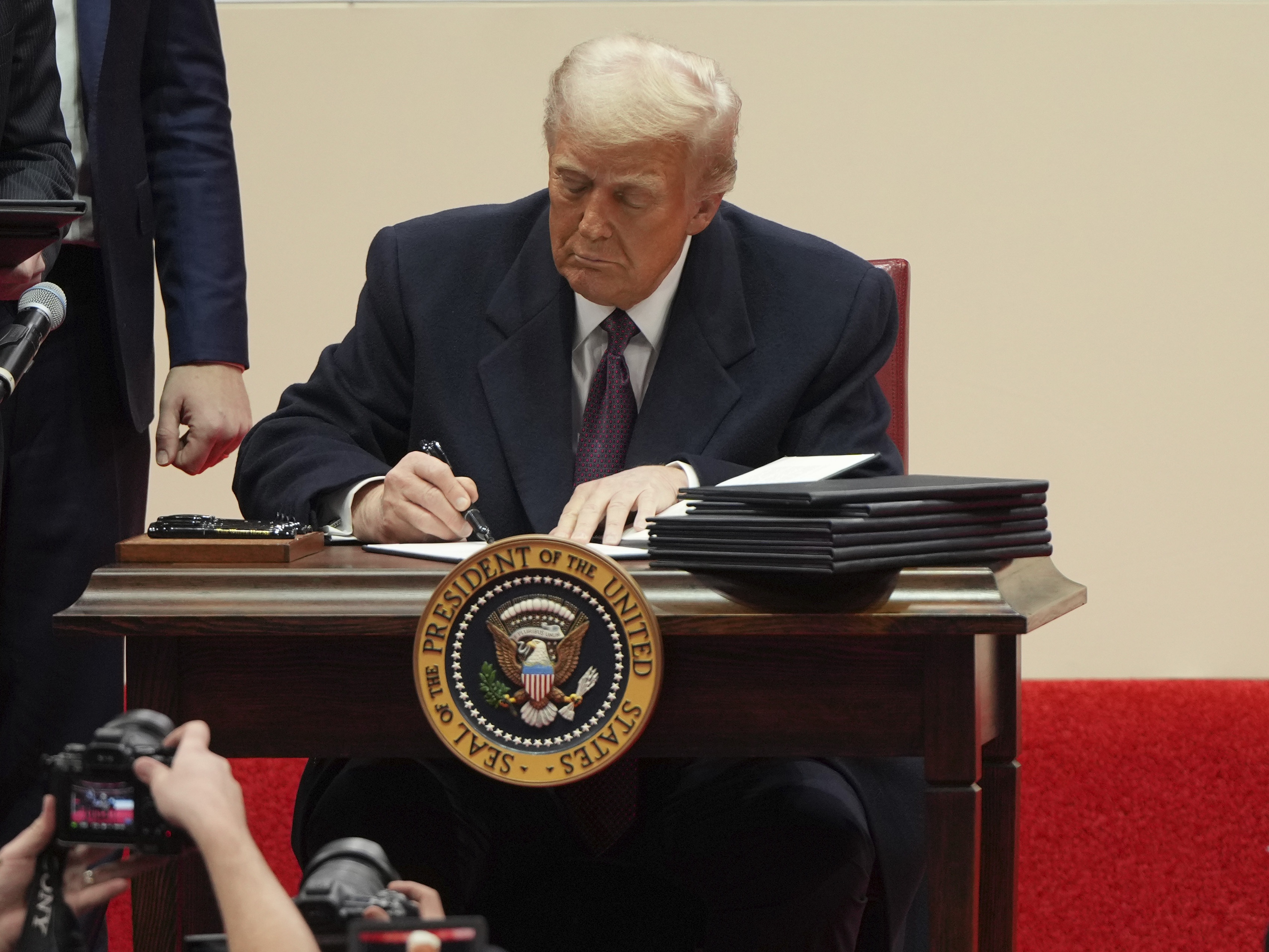 caption: President Donald Trump signs an executive order at an indoor Presidential Inauguration parade event in Washington, Jan. 20, 2025.
