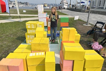 caption: Mila Fedorchenko's ideal city, as she built it during the 2023 Seattle Design Festival. The boxes represent different building types. She stacked some on top of each other to make more room for parks. She wants you to know there should be an ice cream shop near every school.