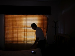 caption: Claudio Rojas, who was deported from the U.S. in 2019, is silhouetted against a curtained window of his home in Moreno, Argentina, on May 8, 2021. He was able to return to the U.S. in August.