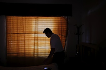 caption: Claudio Rojas, who was deported from the U.S. in 2019, is silhouetted against a curtained window of his home in Moreno, Argentina, on May 8, 2021. He was able to return to the U.S. in August.