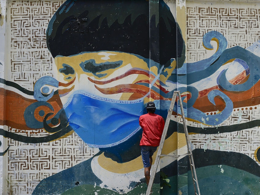 caption: A street artist spray-paints a protective face mask over an old mural featuring a Venezuelan Indigenous man in Caracas, Venezuela, on Saturday. Globally, new daily cases hit an all-time high on Saturday, the World Health Organization reports.
