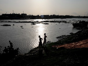 caption: Two boys stand at the edge of the Buriganga River in Dhaka, Bangladesh, in July. A recent study finds that globally, boys and young men made up two-thirds of all deaths among young people in 2019.