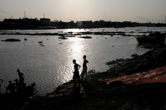caption: Two boys stand at the edge of the Buriganga River in Dhaka, Bangladesh, in July. A recent study finds that globally, boys and young men made up two-thirds of all deaths among young people in 2019.