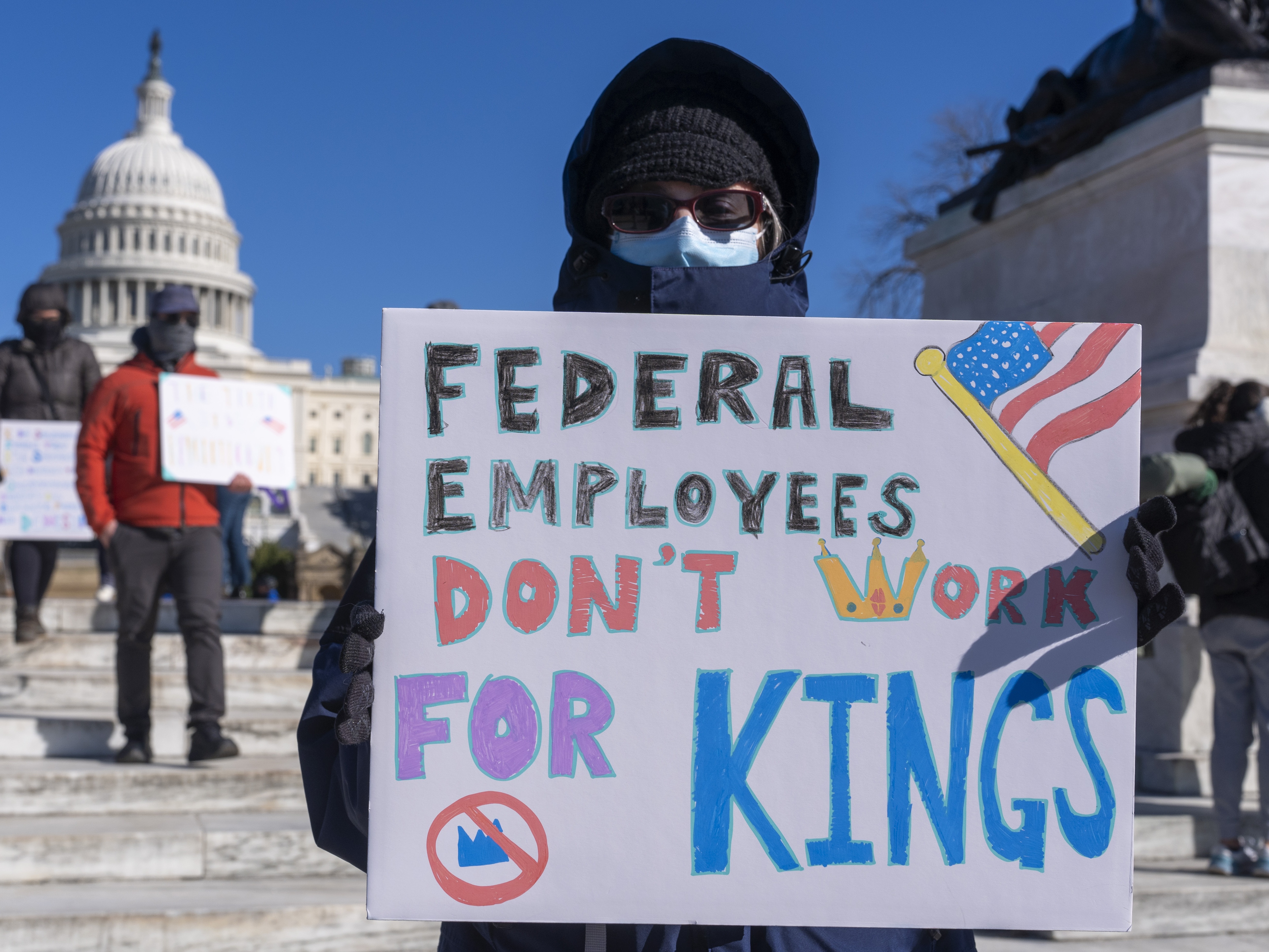 caption: A protester with a sign saying "Federal Employees Don't Work for Kings" demonstrates in support of federal workers and against recent actions by President Trump and Elon Musk on Presidents Day in Washington, D.C.