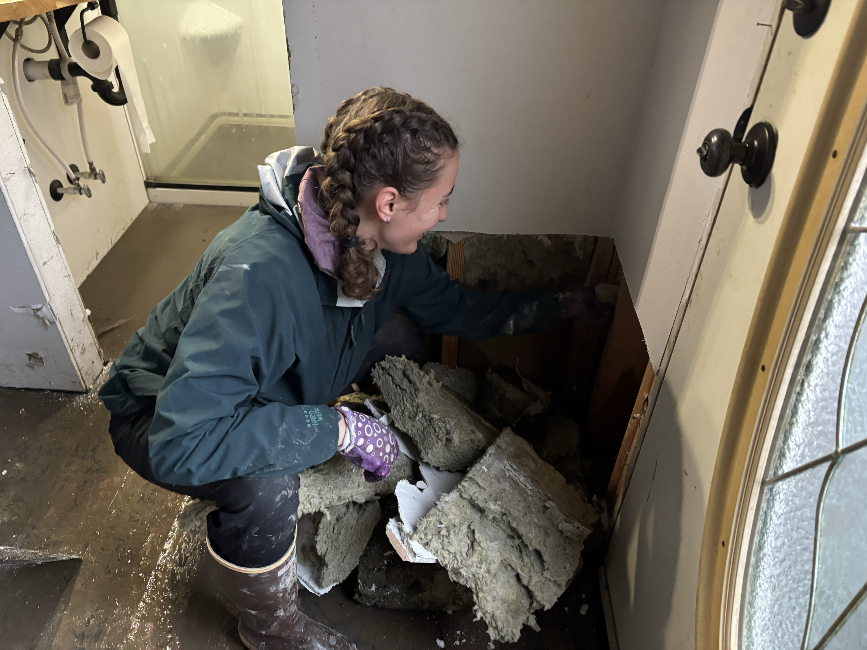 caption: Lauren Karp, a volunteer who drove in from Bellingham, Wash., pulls soggy insulation out of the walls in a muddy home in Sumas, Wash., on Dec. 13, 2025.