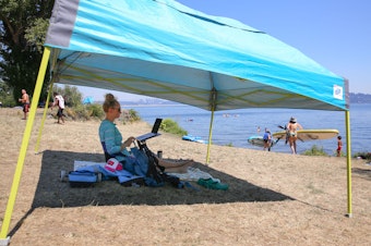 caption: Jenna Shimek works on her laptop at Colman Beach, Monday, between periodic dips in the Lake Washington. "It's cooler than anywhere else I could be," she said.
